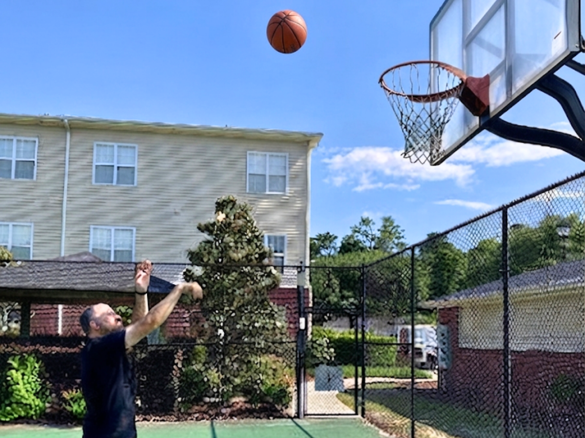 Man on an outdoor basketball court shooting a ball toward the hoop against a blue sky, with apartment buildings in the background.
