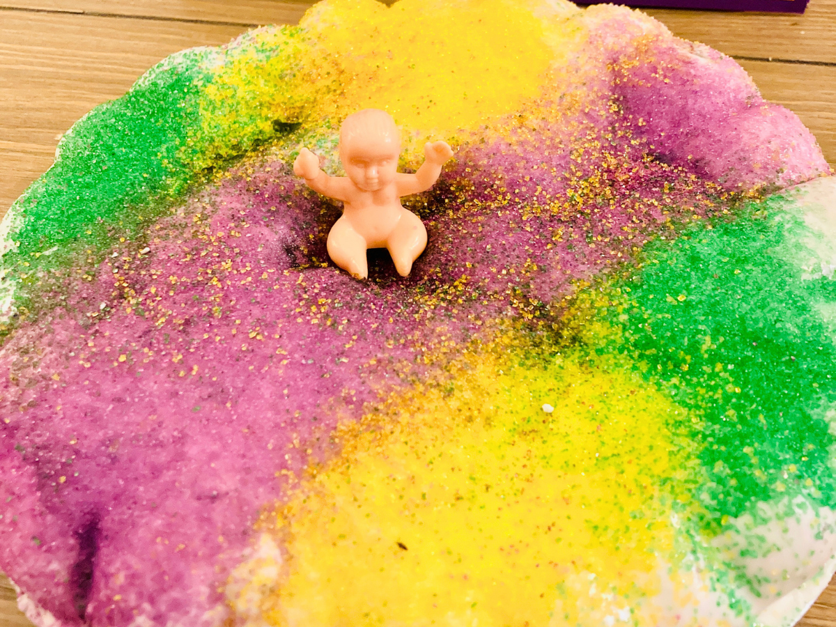 Plastic baby figurine sitting atop a rainbow-colored sugar dish on a plate, with green, yellow, purple, and pink granules surrounding it.