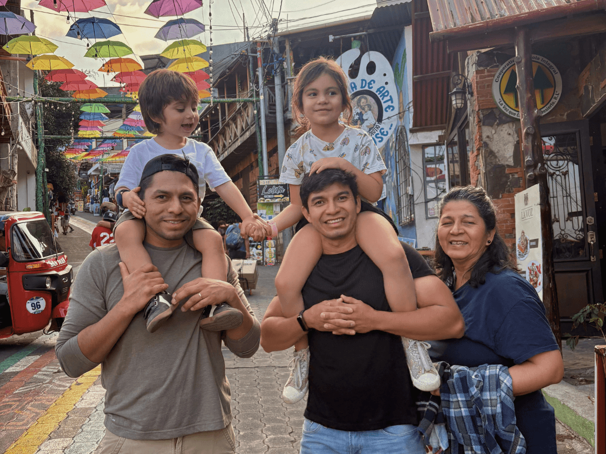 Family of four posing on a colorful street with rainbow umbrellas overhead; two dads carry kids on their shoulders, mom beside them.
