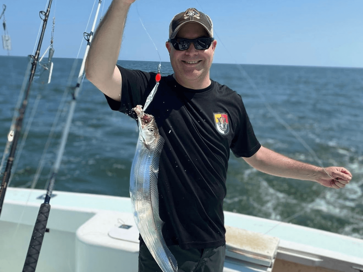 Man on a boat wearing sunglasses and a black shirt holds up a long, silvery ribbonfish he just caught while fishing at sea.