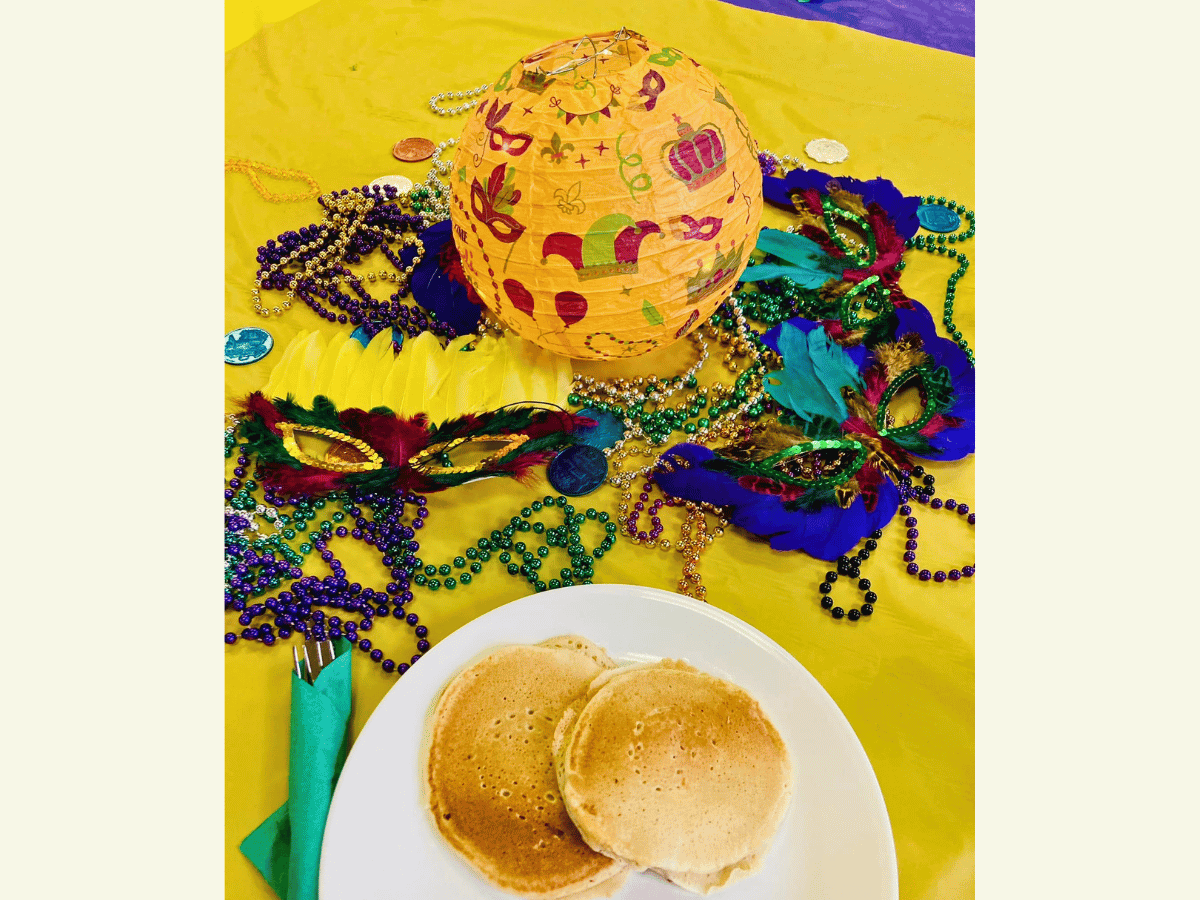 Plate with two pancakes on a yellow table, surrounded by colorful Mardi Gras beads, masks, and a decorative yellow lantern.