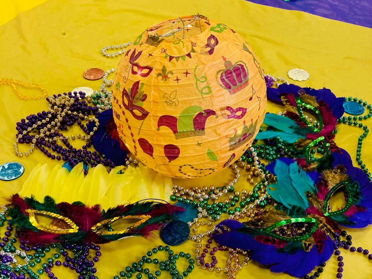 Orange paper lantern decorated with carnival icons, surrounded by colorful beads and masquerade masks on a yellow surface for Mardi Gras.