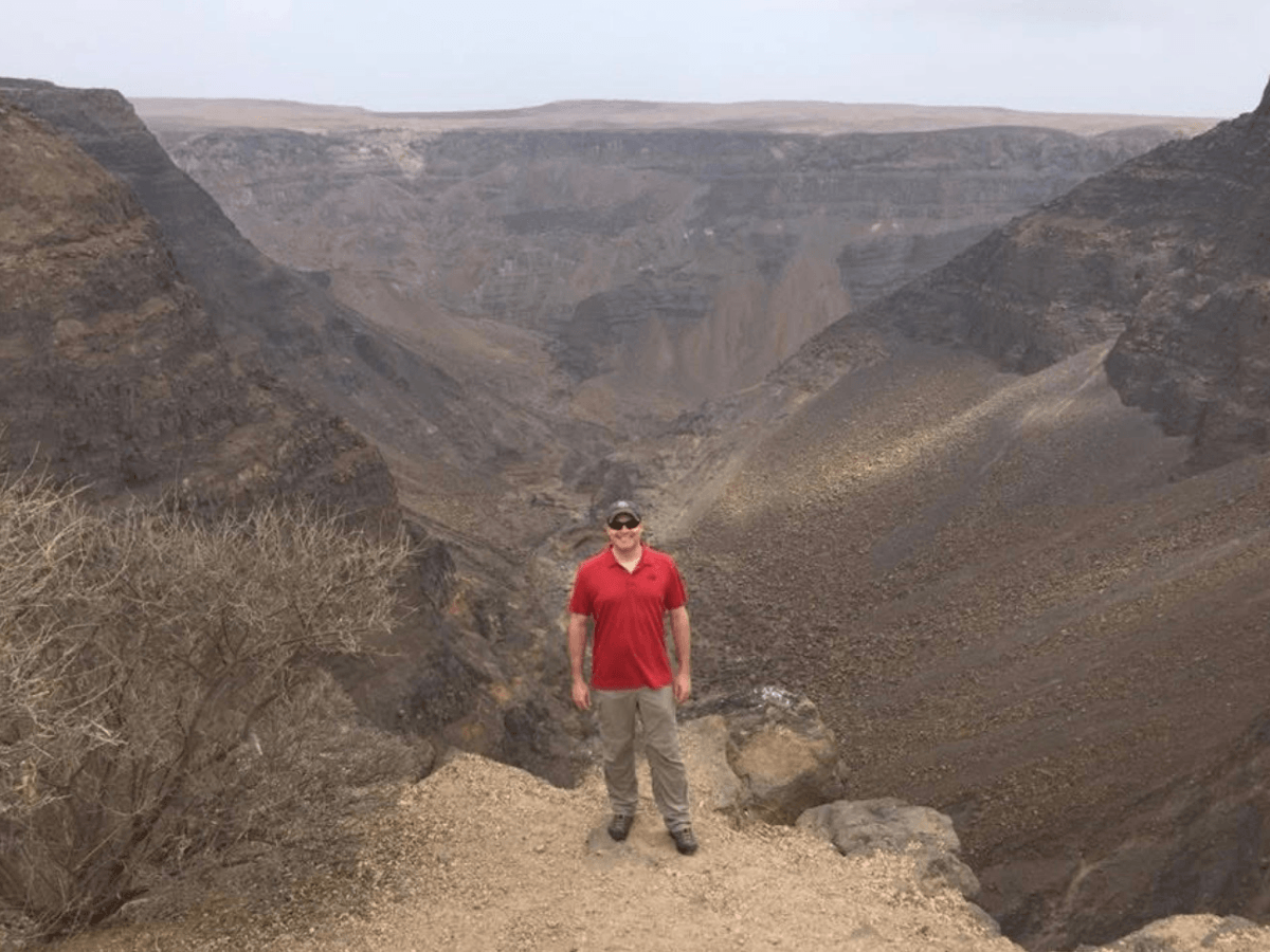 Man in a red shirt and beige pants standing on a rocky canyon overlook with steep cliffs in the background.