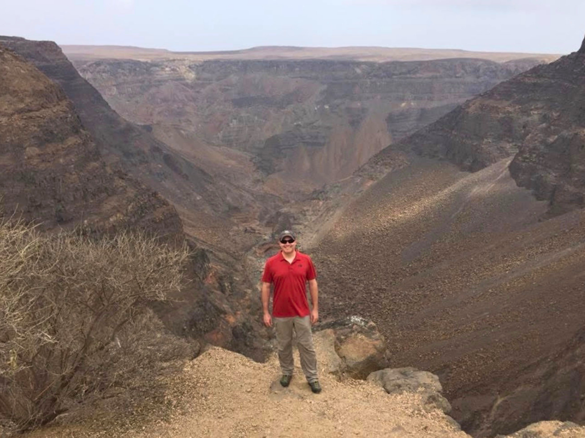 Man in a red polo stands on a rocky canyon rim, overlooking a deep ravine with brown cliffs.