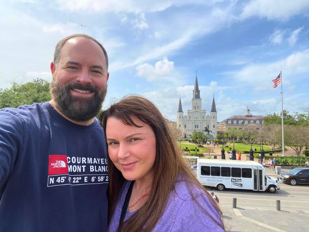 Smiling couple takes a selfie on a sunny day with a cathedral and a white tour bus in the background.