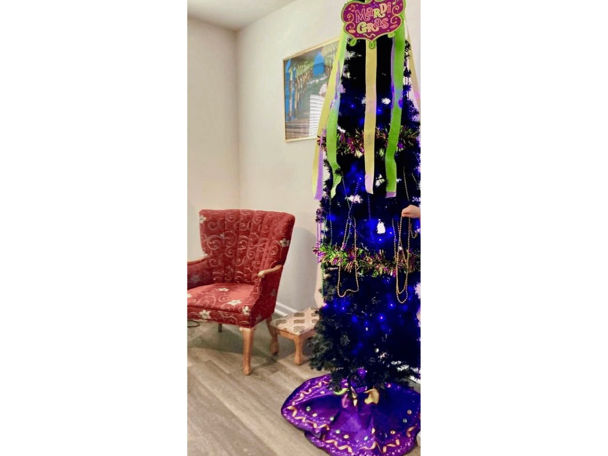 A Christmas/party tree decorated for Mardi Gras with purple lights, green streamers, and bead garlands next to a red upholstered chair in a cozy room.