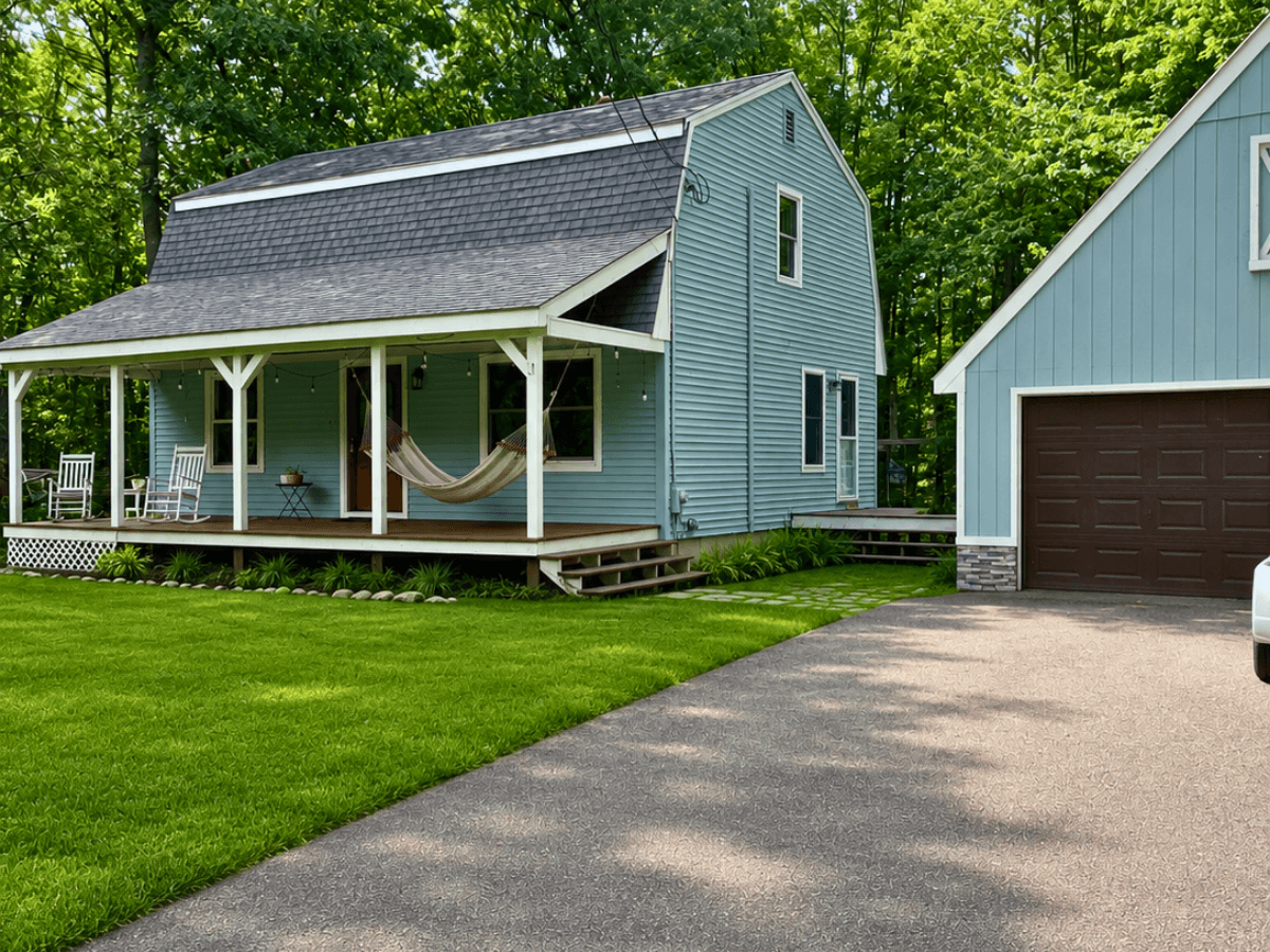 Blue two-story house with a wooden front porch, hammock, rocking chairs, and string lights, next to a grey garage on the right.