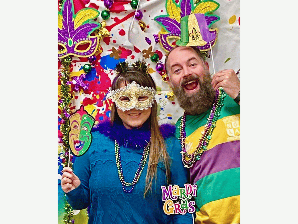 Two adults in Mardi Gras costumes posing with masks and beads against a colorful confetti backdrop.
