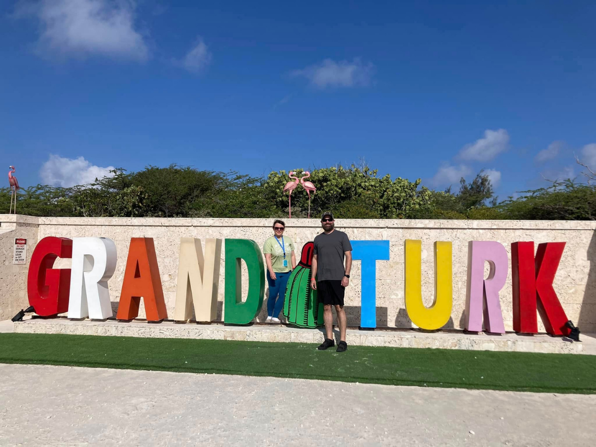 Two people posing in front of a large multicolored 'GRAND TURK' sign against a bright blue sky, with pink flamingos perched on the greenery behind.