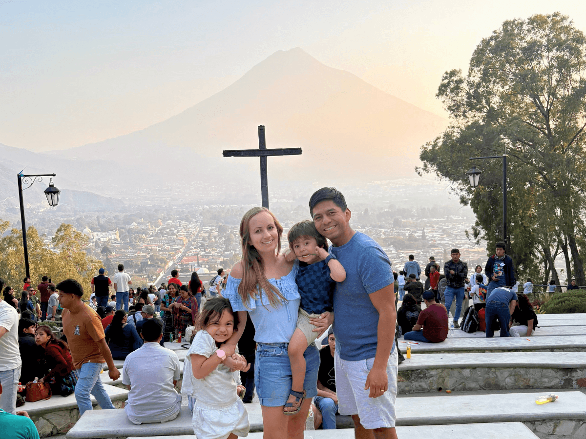 Family posing on steps with a distant volcano and a crowd in the background; a mom, dad, and two children smiling at the camera.