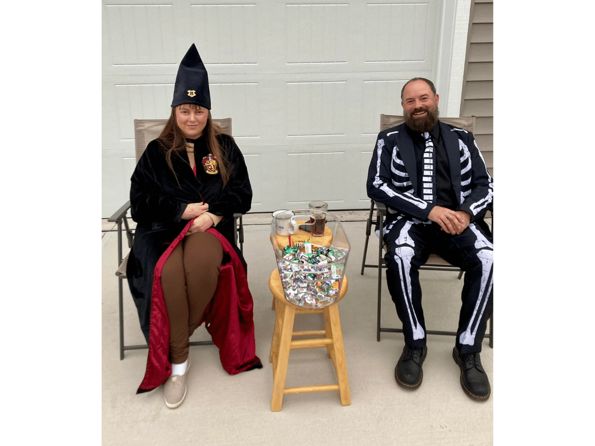 Two people in costumes sit outside a garage; a bowl of candy sits on a wooden stool between them, the left person wearing a wizard-like hat and robe, the right in a skeleton suit with a striped tie over a black shirt.