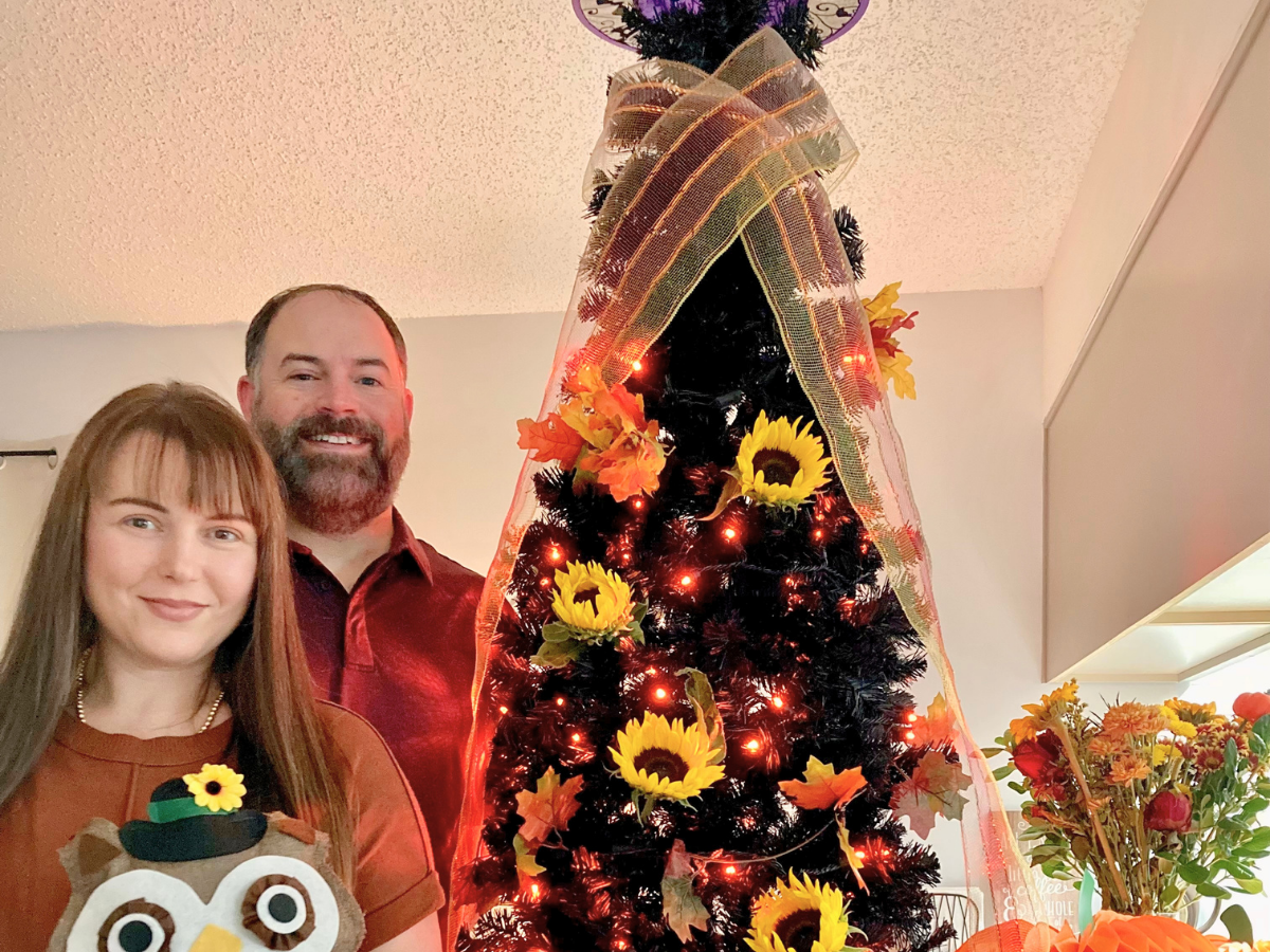 Smiling couple posing indoors beside a tall Christmas tree wrapped in orange ribbon with sunflowers and autumn leaves, lit with red lights.