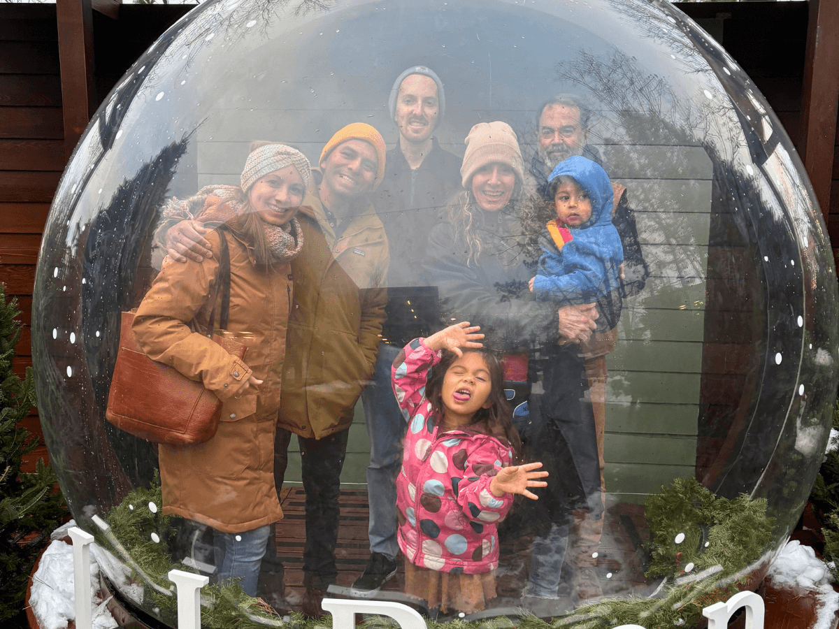 Family of adults and two children posing inside a large clear snow globe outdoors in winter; everyone smiles at the camera, dressed in warm coats and hats.