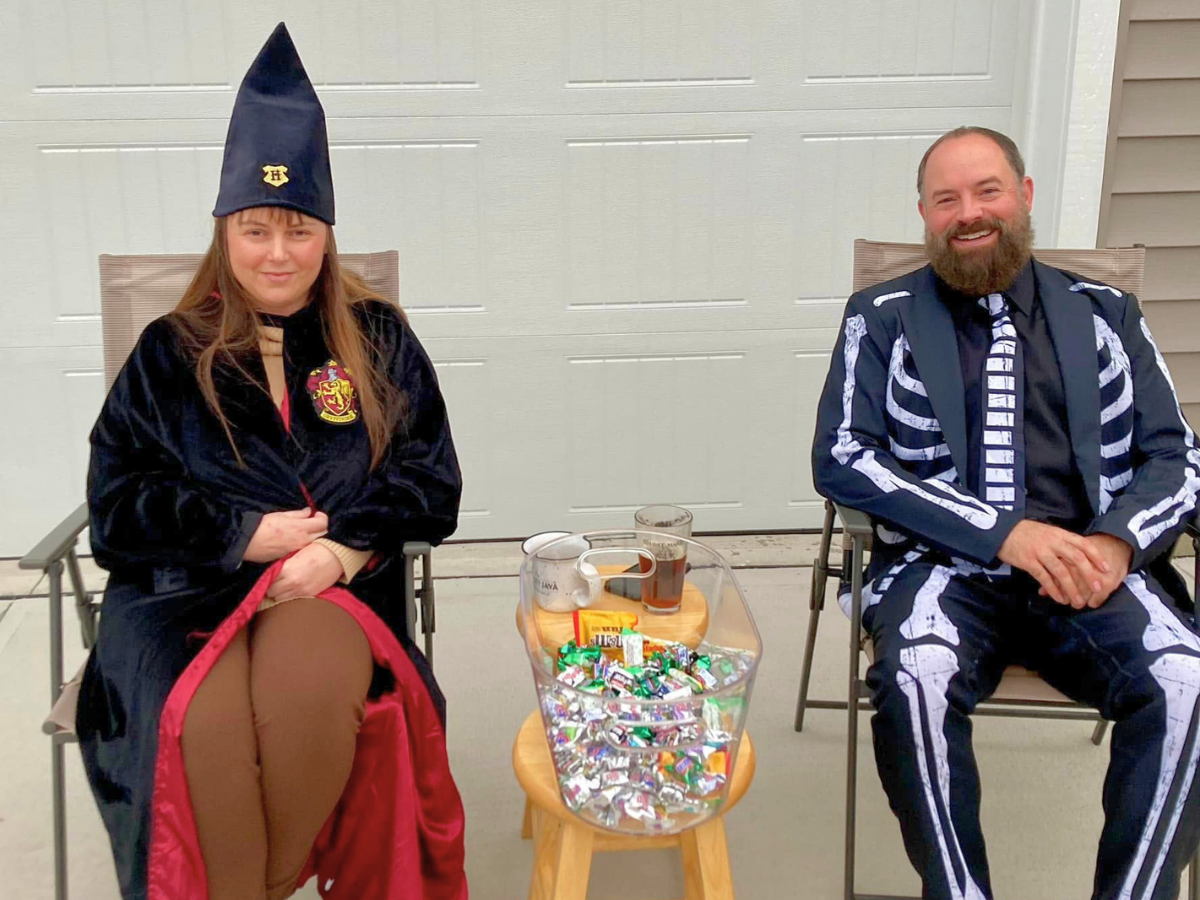 Two people in Halloween costumes sit on chairs outside; woman wears a witch outfit with a tall hat, man wears a skeleton suit, with a candy-filled bowl on a small table between them.