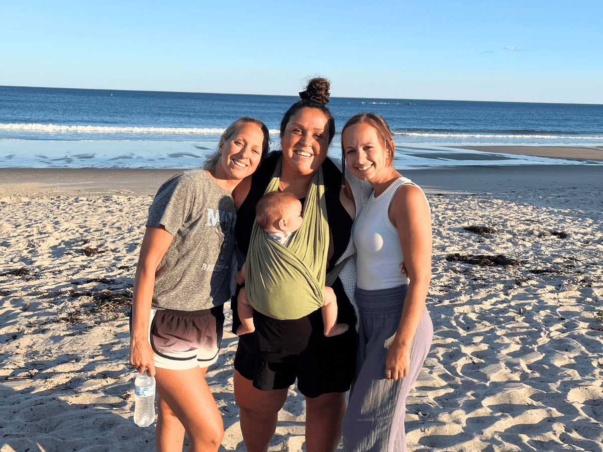Three women on a sunny beach smiling, with a baby in a green wrap carried by the center person, ocean and waves in the background.