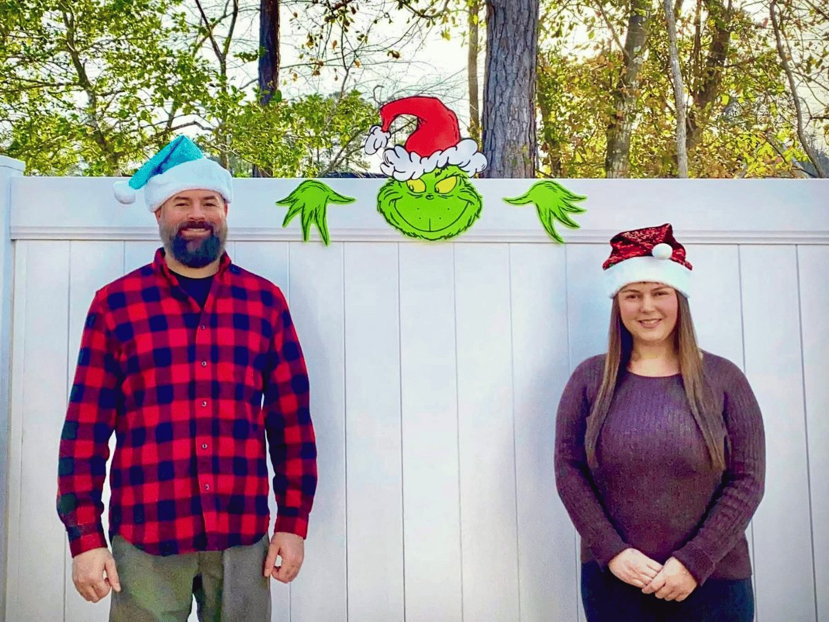 Two adults wearing Santa hats stand in front of a white fence with a Grinch cutout between them.