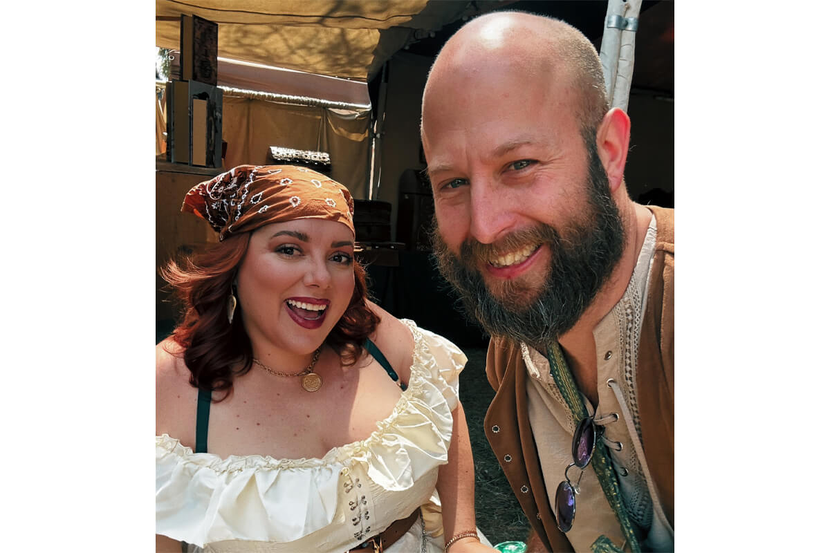 Smiling couple posing for a selfie at a historical fair; woman in a cream off-shoulder top with a bandana, man with a beard in brown vest and laced shirt.