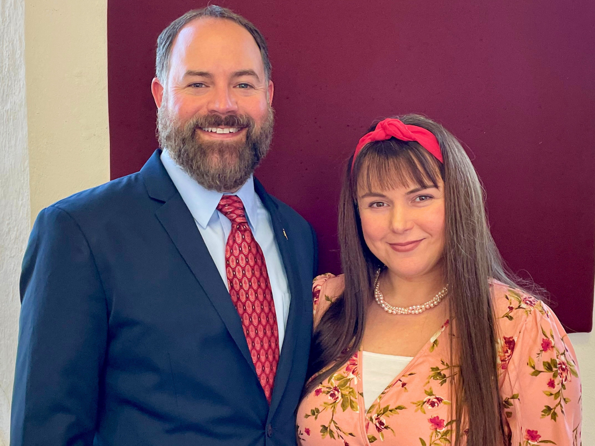 Smiling man in a navy suit and red tie stands beside a woman in a pink floral top with a red headband against a maroon wall.