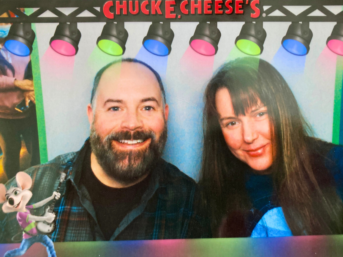Smiling man with a beard and a woman pose for a selfie at Chuck E. Cheese's, with colorful overhead lights in the background and a cartoon mouse on the left.