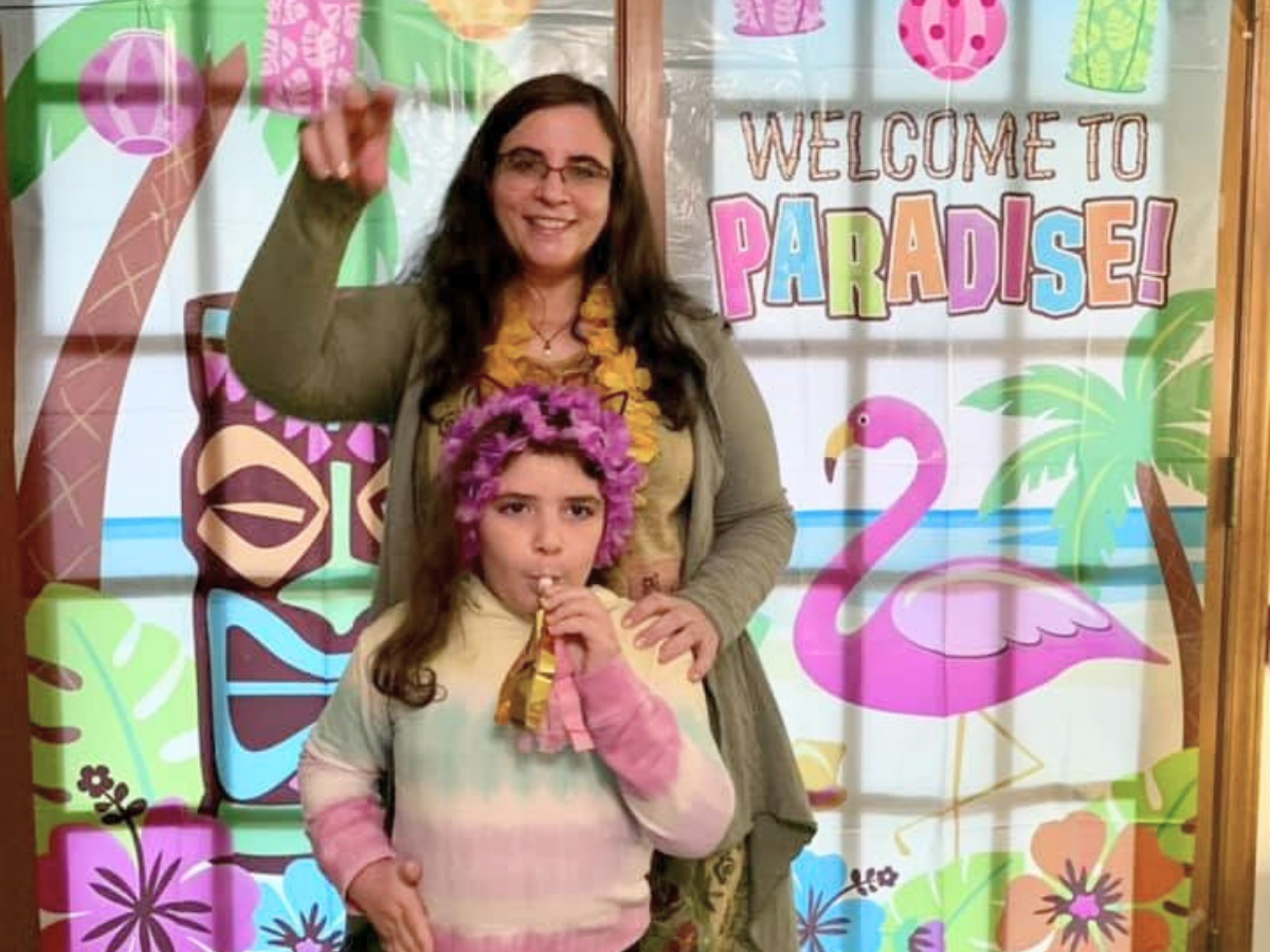 Woman standing behind a girl in a pink tie-dye sweater, both posing in front of a colorful 'Welcome to Paradise' backdrop.