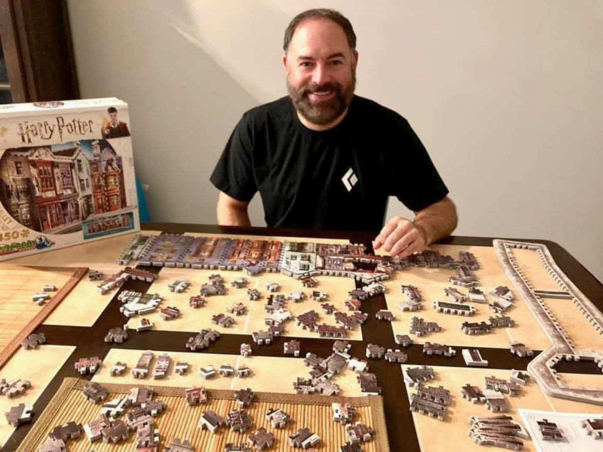 Man with a beard in a black shirt sits at a table playing a large tile-based board game with many scattered pieces and a boxed set on the left.