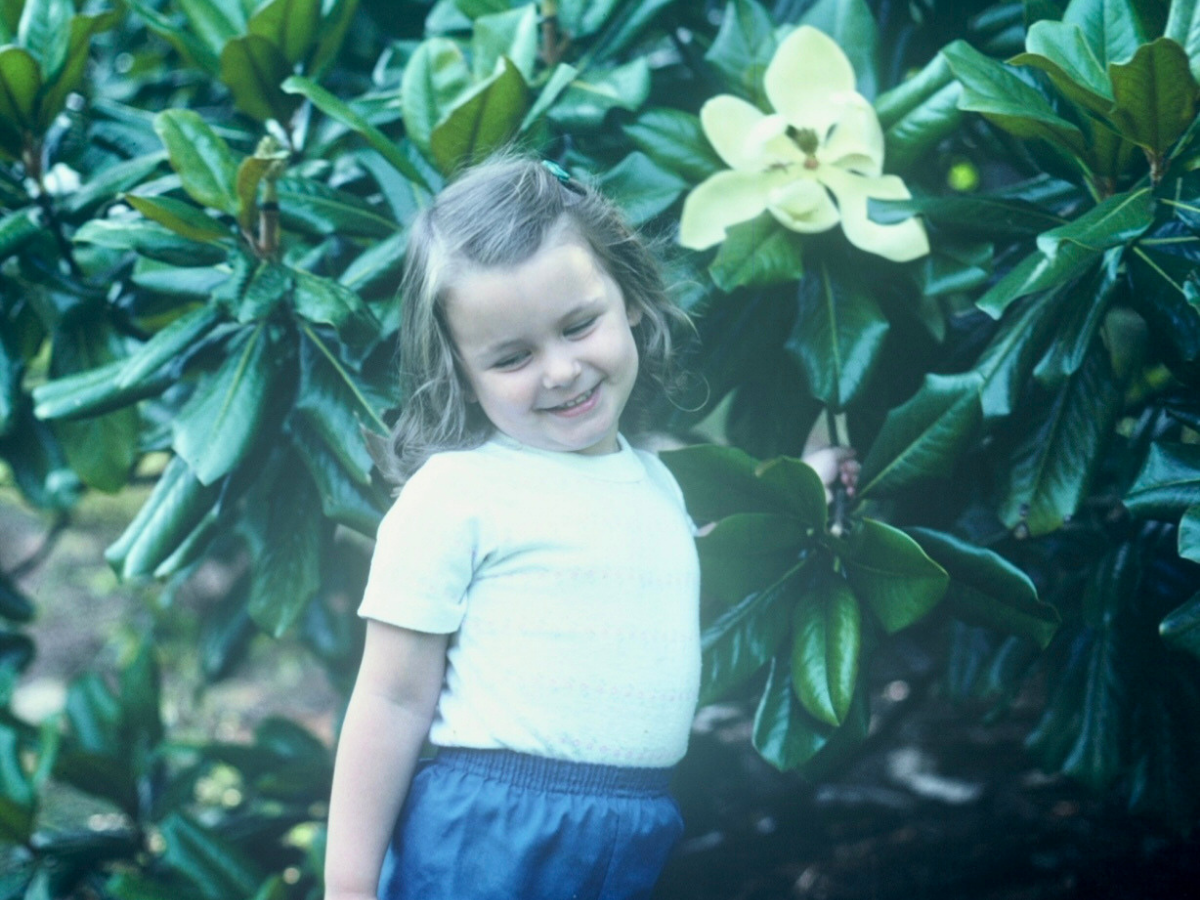 Smiling young girl in a white shirt and blue shorts stands among glossy green leaves with a pale yellow flower nearby.