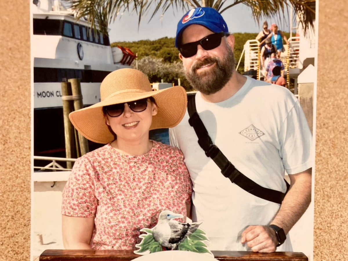 A smiling couple posing together outdoors, woman wearing a wide straw hat and glasses, man in a light shirt and blue cap with sunglasses.