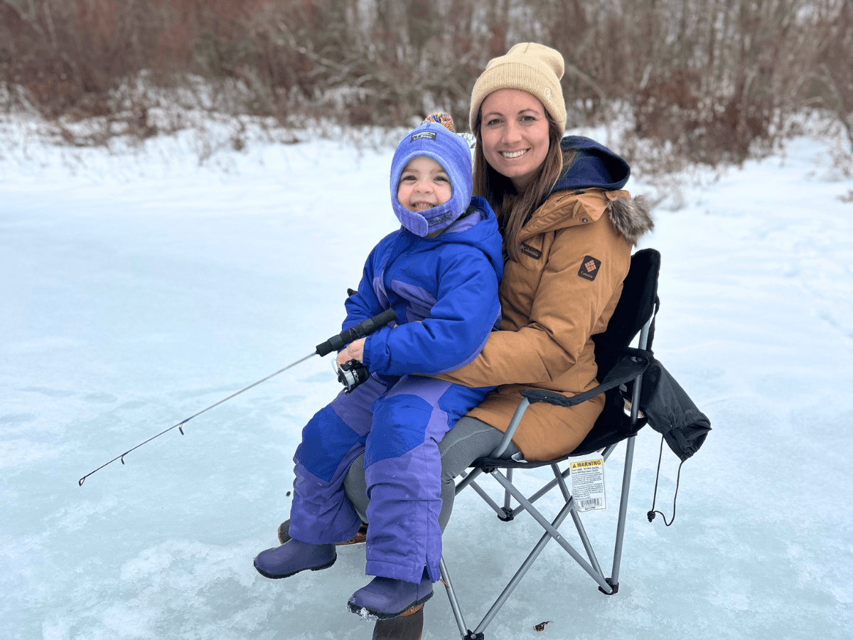 Mother and child in winter jackets sit on a folding chair on a frozen lake, fishing together.