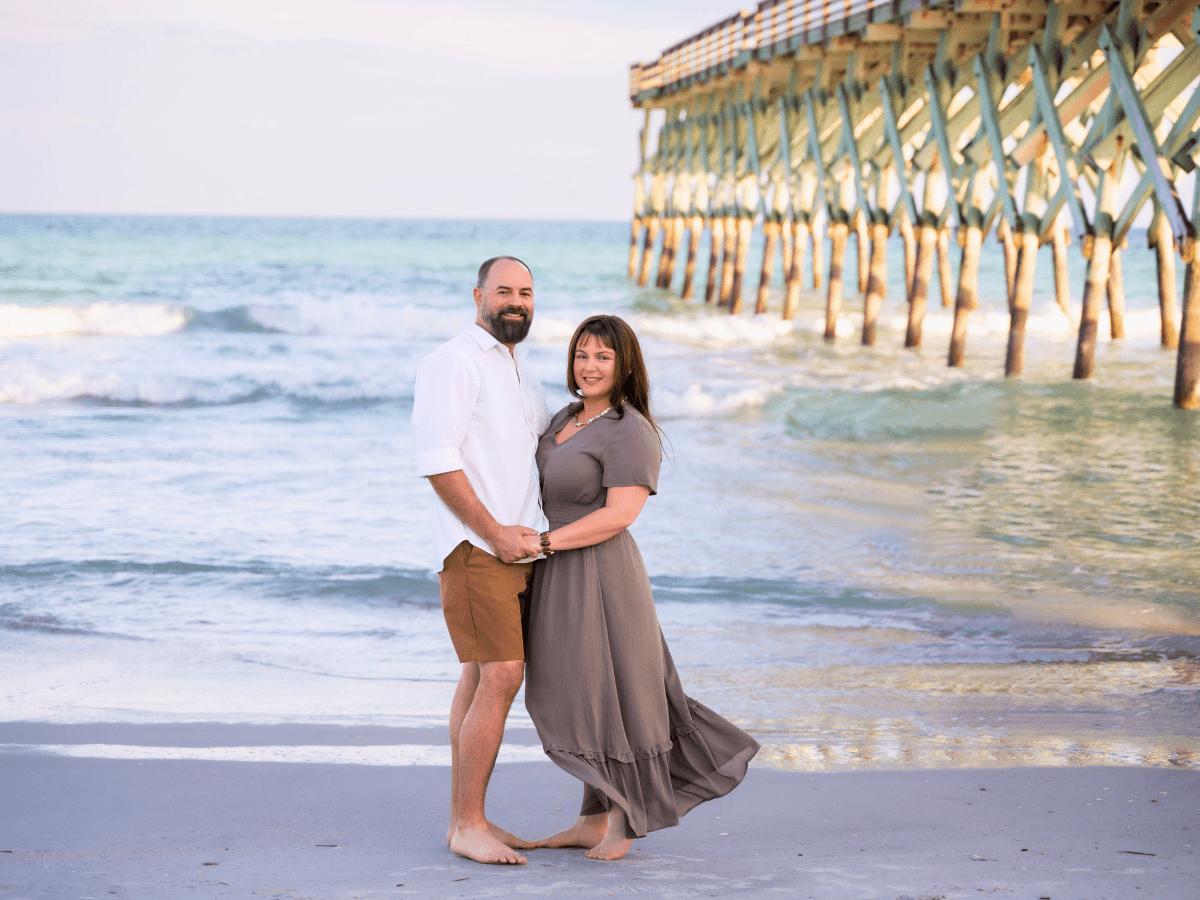 Couple stands on a sandy beach by a wooden pier, holding hands and smiling at the camera.