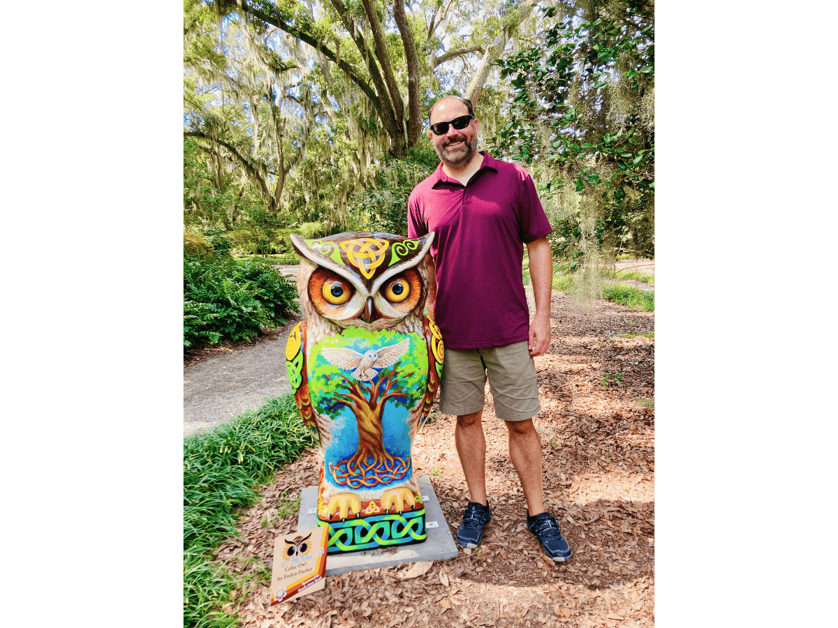 Man in a maroon polo and shorts posing next to a brightly painted owl sculpture in a wooded park.