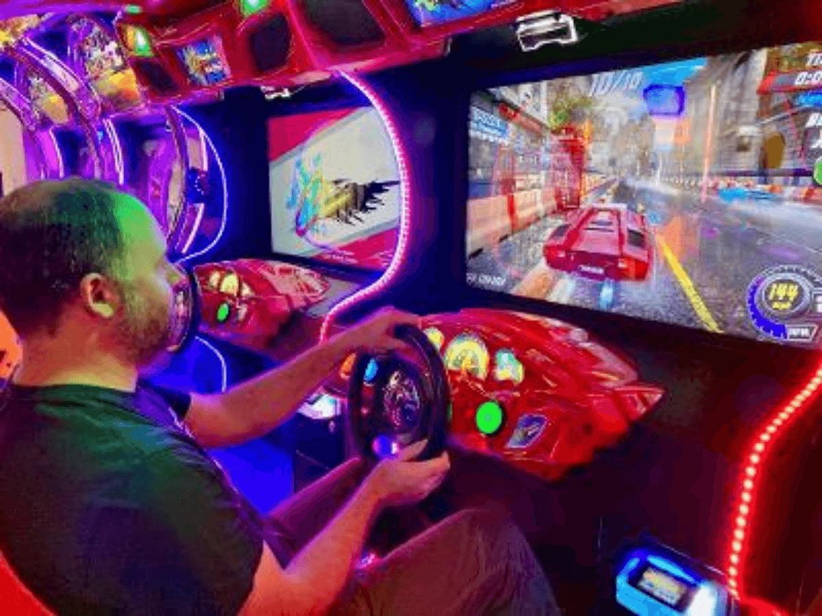 Man in a black shirt seated in a neon-lit arcade racing cabinet, steering a colorful game screen ahead of him to race virtual cars.