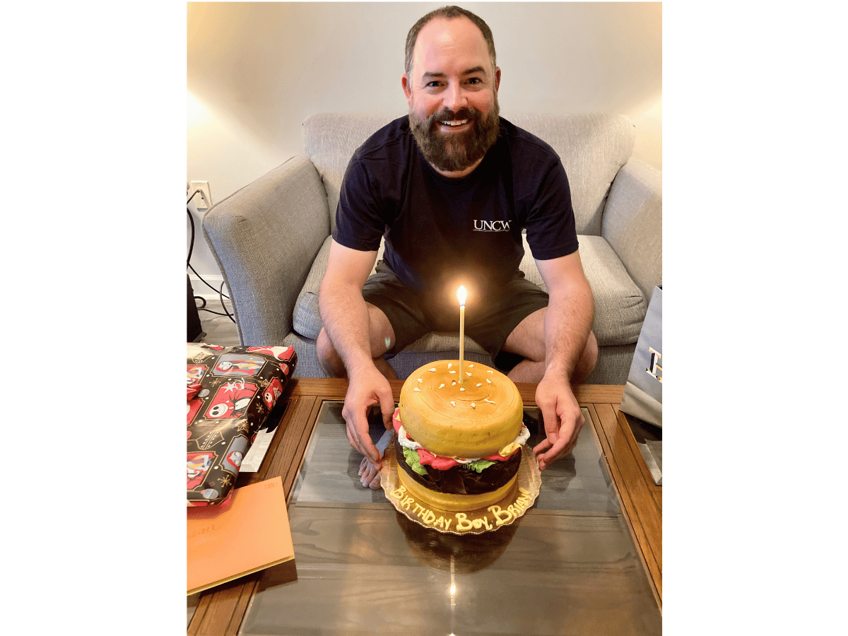 Smiling man with a beard sits on a gray sofa, birthday cake shaped like a hamburger with one lit candle in front of him.