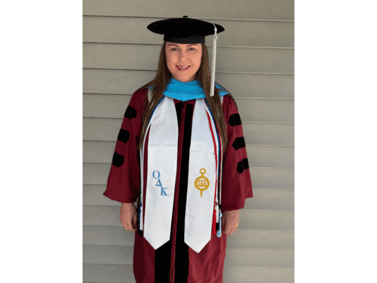 Graduate in maroon gown and black mortarboard, with a white stole bearing blue letters and a gold emblem, standing against a siding wall.