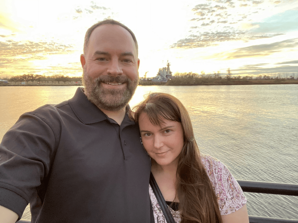 Smiling couple taking a sunset selfie by a calm river, railing in the foreground with a distant ship on the horizon.