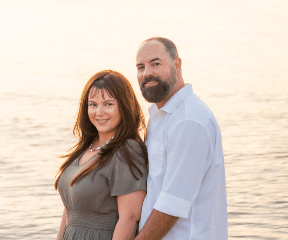 Smiling couple standing side by side by a calm body of water at sunset, the man in a white shirt and the woman in a gray dress.
