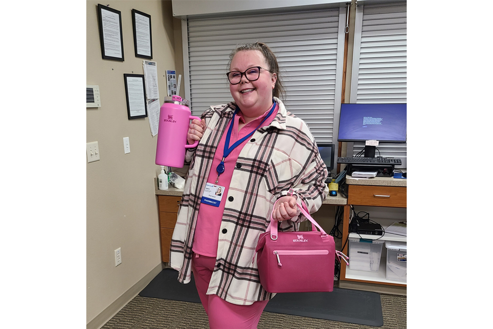 Smiling woman in a pink shirt and plaid coat, wearing a name badge, holds a pink water bottle and a pink bag in an office setting.