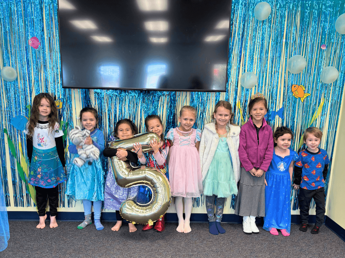 Group of smiling children in colorful costumes posing for a photo at a party, holding a large gold number 5 balloon in front of a blue fringe backdrop.