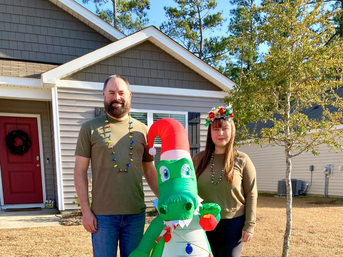 Man and woman stand in a front yard with a large green inflatable alligator wearing a red Santa hat, in front of a grey house with a red door.