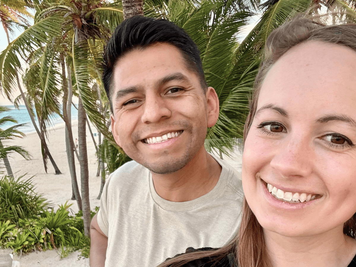 Smiling couple posing for a selfie on a sandy beach with palm trees and ocean in the background.