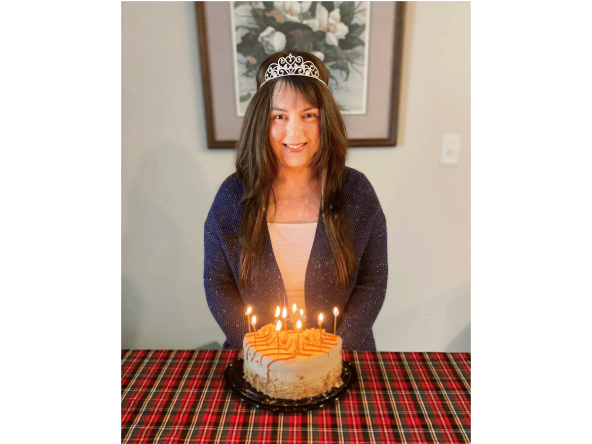 Young woman wearing a tiara sits at a birthday cake with lit candles on a plaid tablecloth from the waist up, smiling at the camera.