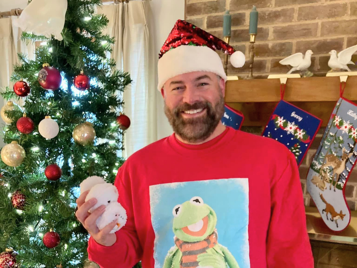 Man in a Santa hat and red sweater featuring Kermit the Frog, smiling and holding a snowball in front of a Christmas tree.
