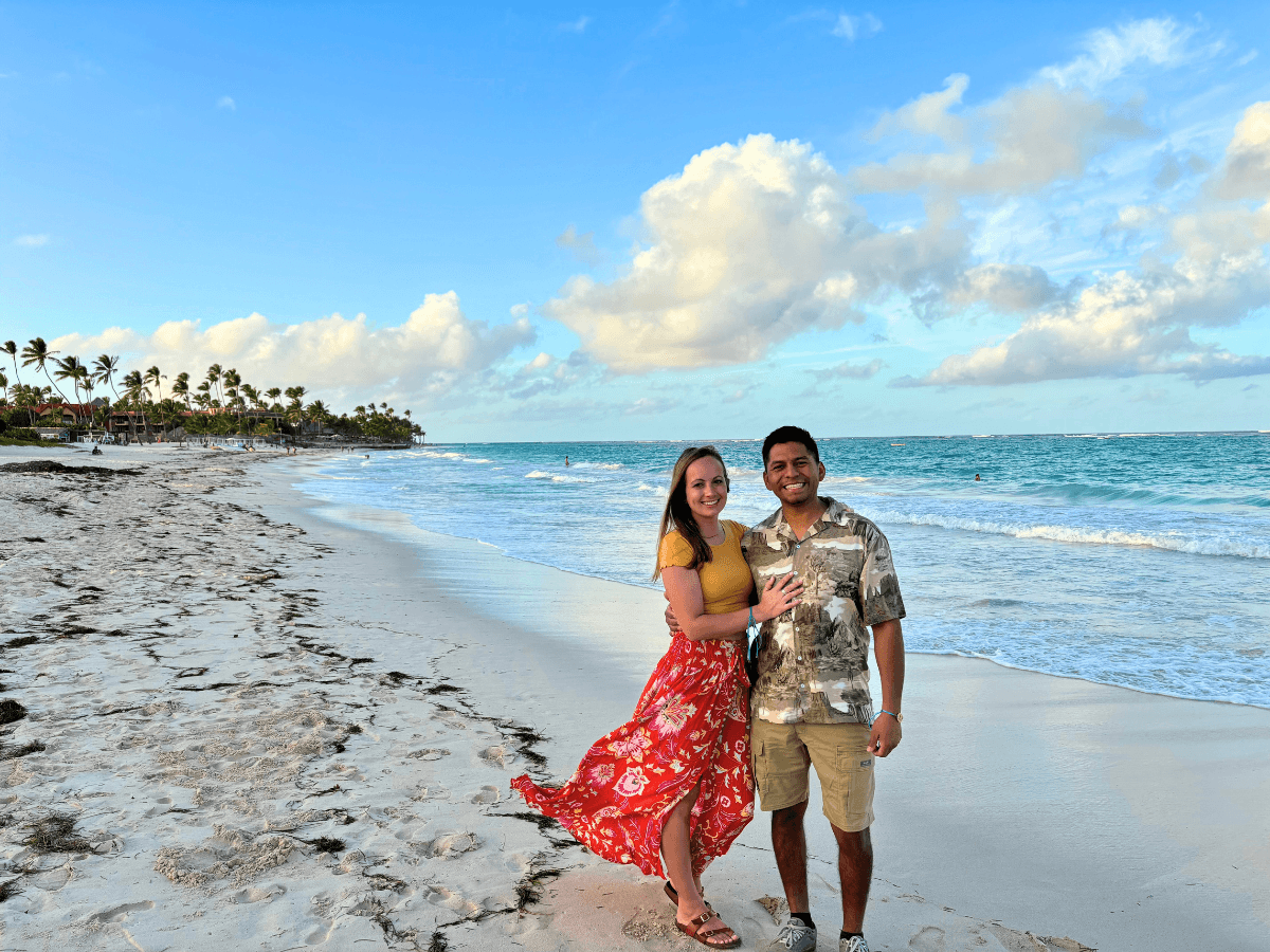 Couple posing on a sunny beach with turquoise water and palm trees in the distance, smiling at the camera in tropical setting short skirt.