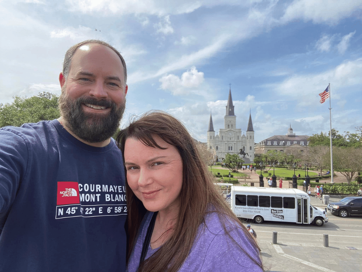 Smiling couple taking a selfie in front of a large cathedral with three spires and an American flag nearby.