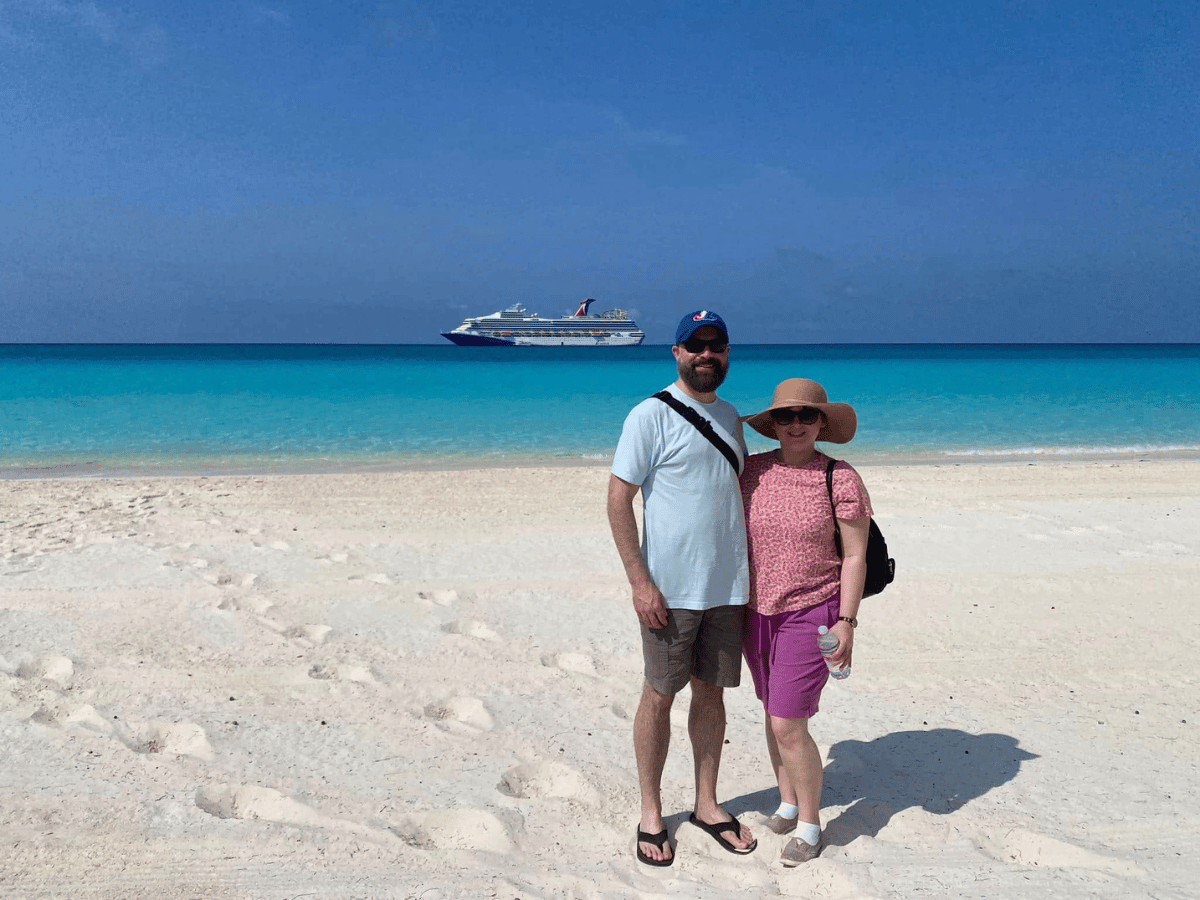 Couple standing on white beach with turquoise sea and a cruise ship in the distance.