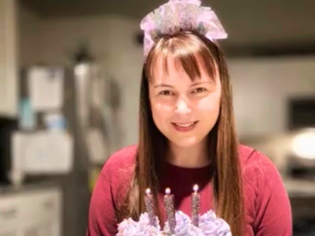 Woman smiling in a kitchen, wearing a pink headpiece, with a frosted birthday cake and lit candles in front of her.