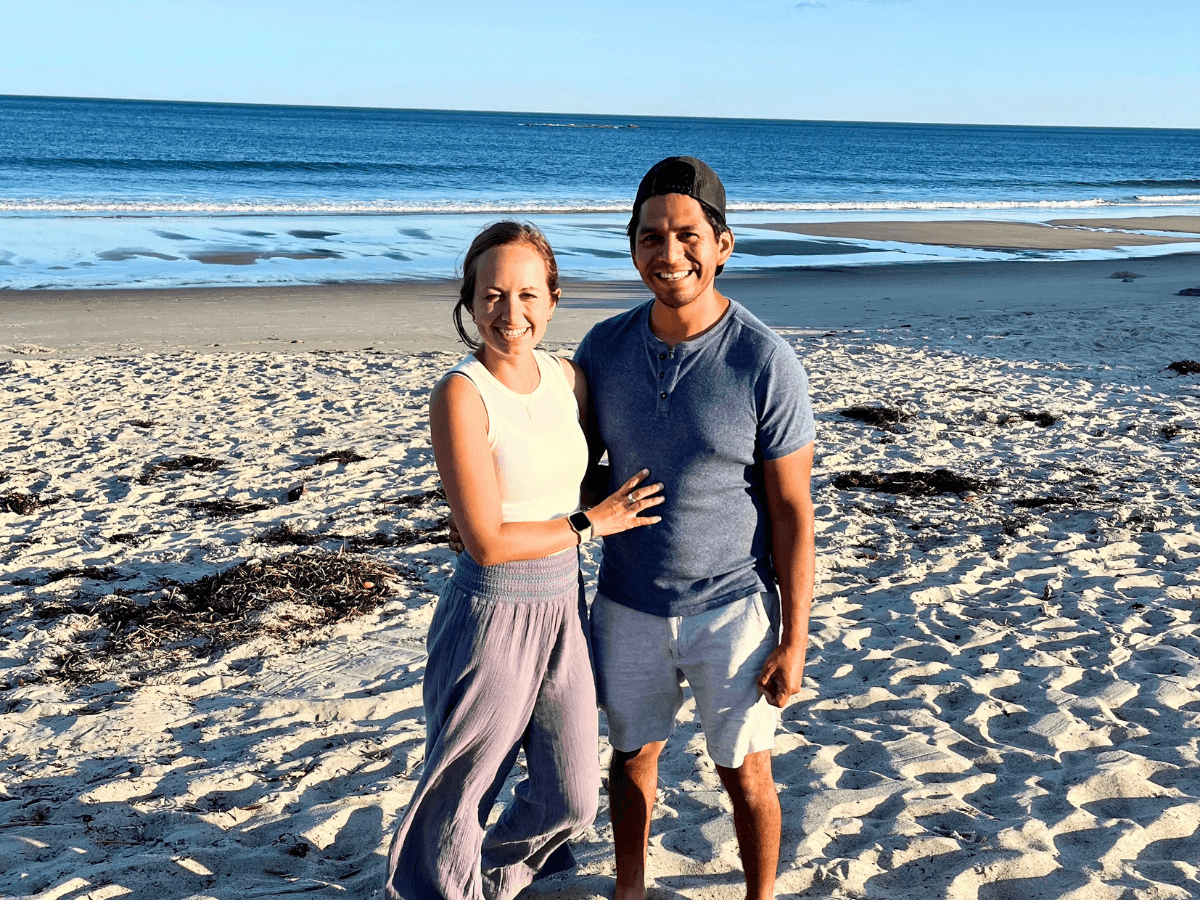 Smiling couple standing arm-in-arm on a sunny beach with the ocean in the background.
