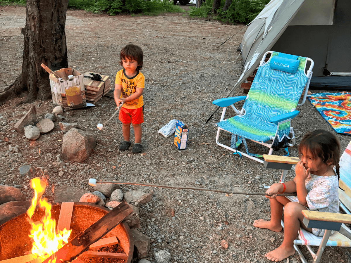 Two children roasting marshmallows over a campfire beside a tent and a blue striped camping chair in a wooded campsite.