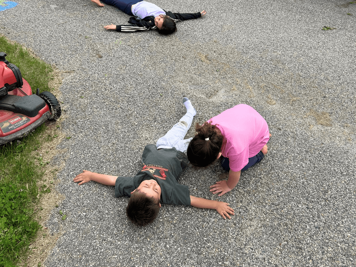 Three children on a gravel driveway; a boy lies on his back in a green shirt while two girls bend over him, with a red lawn mower on the left edge of the scene.