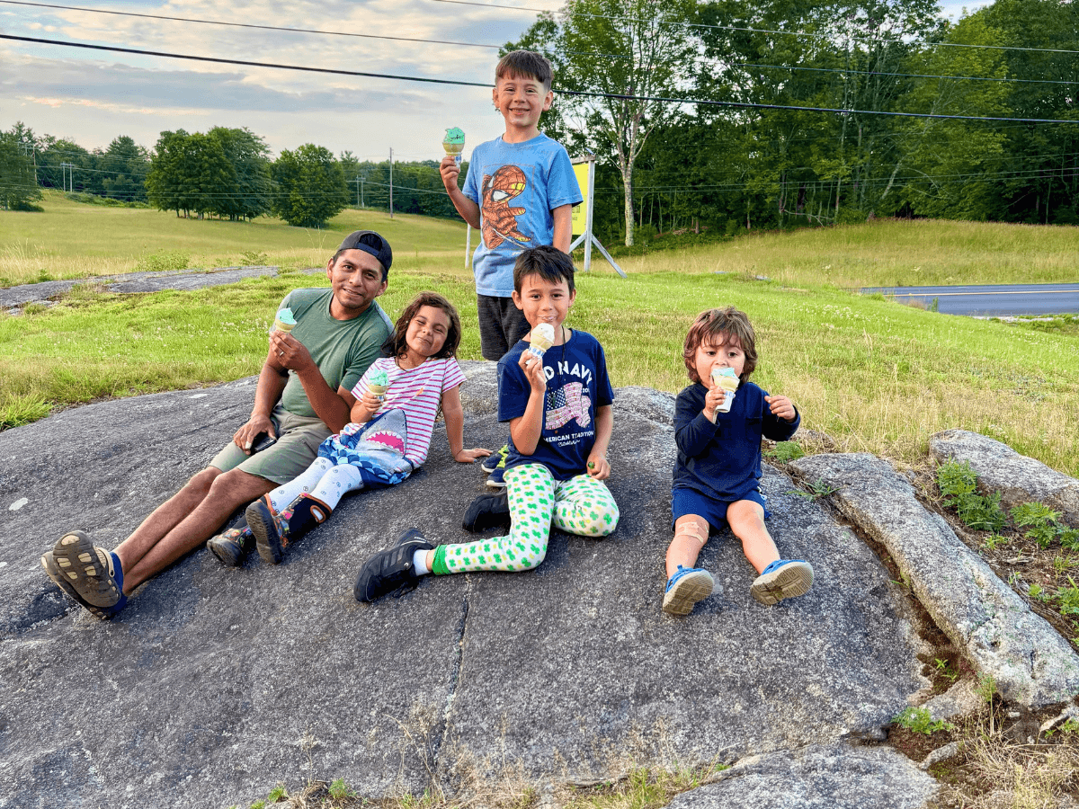 Group of five kids and one adult sitting on a large rock outdoors, each holding ice cream cones.