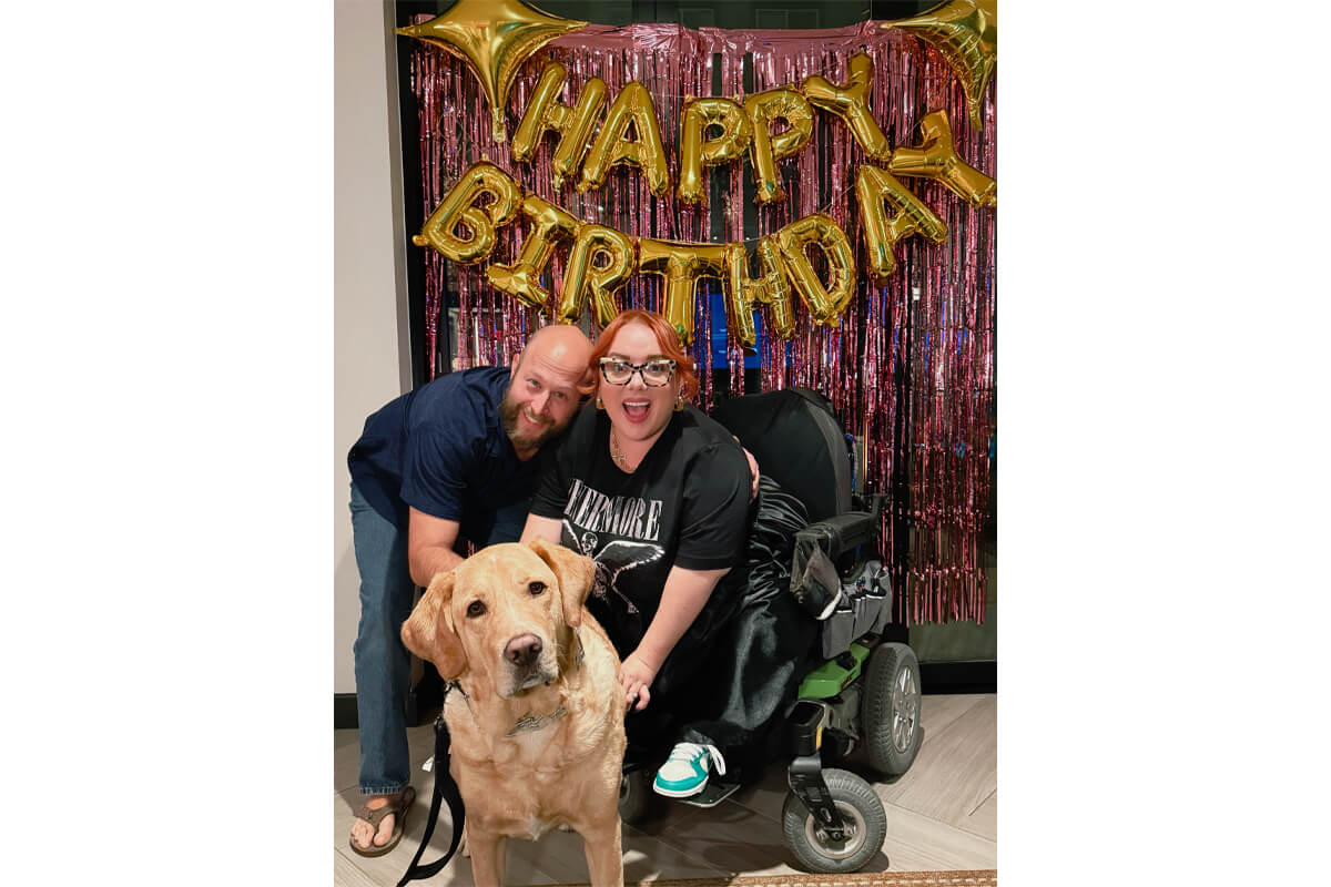 Man and woman smile with their dog in front of a gold 'Happy Birthday' balloon banner and pink fringe backdrop; woman sits in a motorized wheelchair.